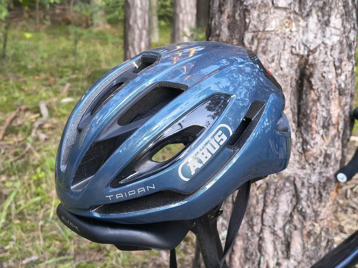 Kask rowerowy założony na siodełko rowerowe. Dobrze widziana nazwa i logo producenta: ABUS Taipan.