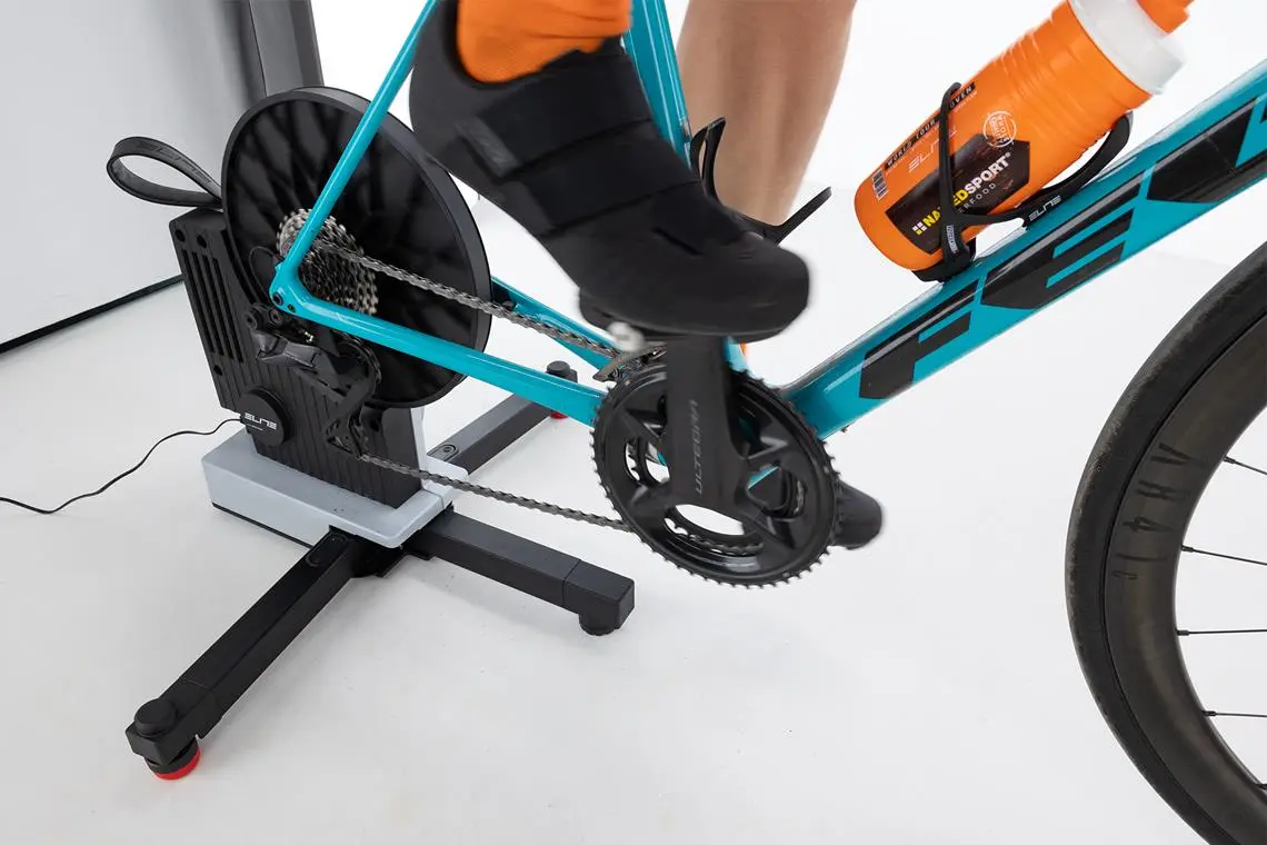 Trenażer rowerowy marki Elite z zamocowanym rowerem Felt, na którym pedałuje rowerzysta. W koszyku bidon.