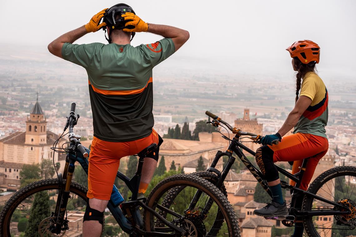 Para rowerzystów MTB stoi na wzgórzu nad zabytkowym miastem i patrzy w dal, mężczyzna dopasowuje w tym czasie kask.