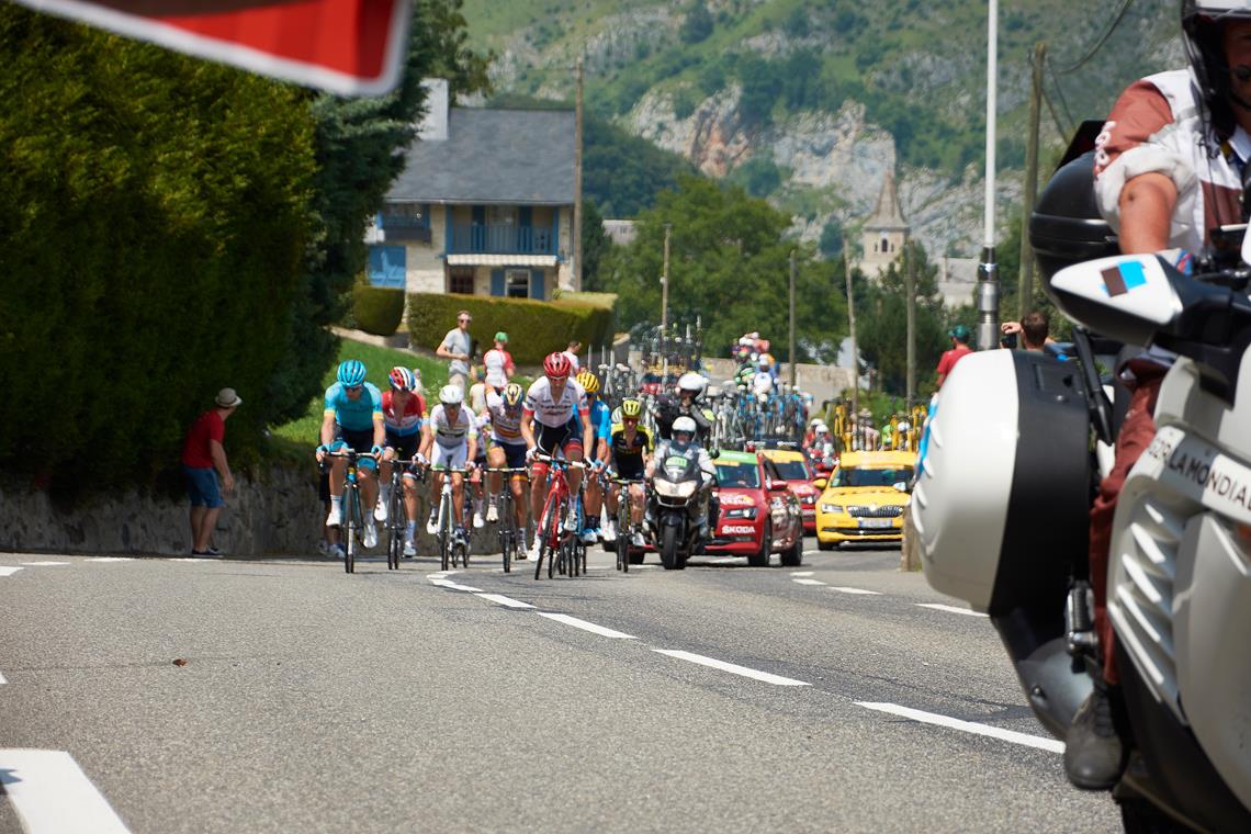 Kolarze w peletonie, za nimi jadą samochody