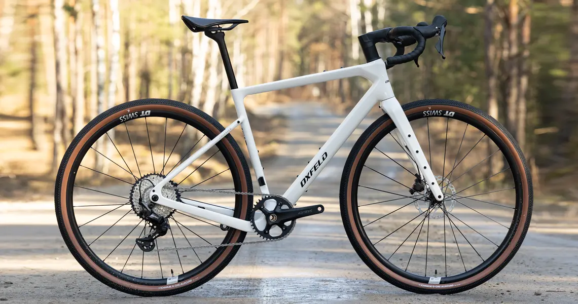 Biały rower gravelowy z karbonu na leśnej ścieżce w słoneczny dzień.