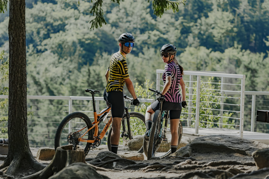 Para rowerzystów stoi w punkcie widokowym w górach razem ze swoimi rowerami.